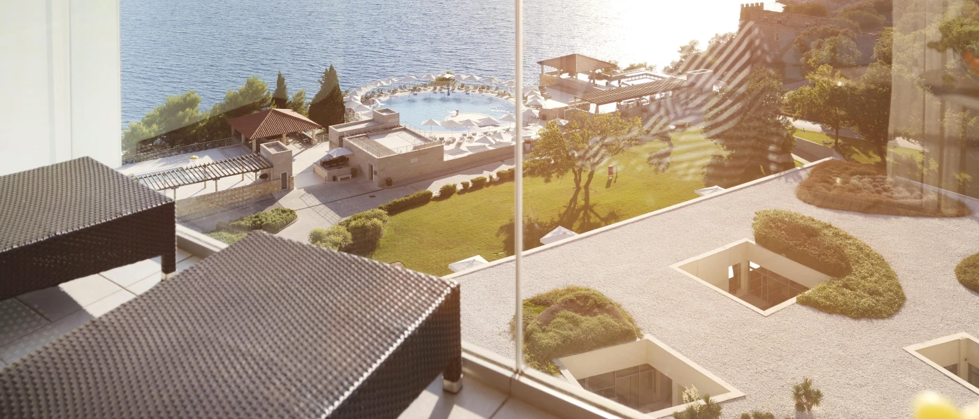 Glass balcony with black rattan loungers, overlooking landscaped grounds, a swimming pool and the ocean below