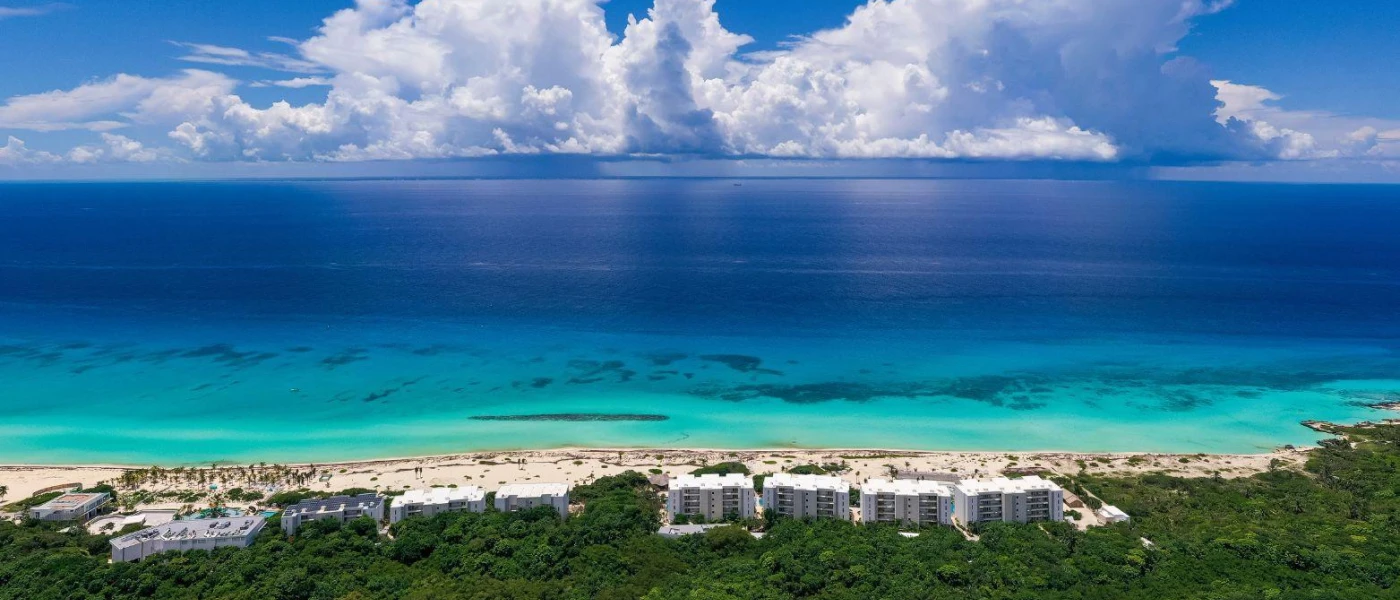 Thick green forest next to a high-rise white resort, with white sands and turquoise shallows in front of it, under a blue and cloud sky