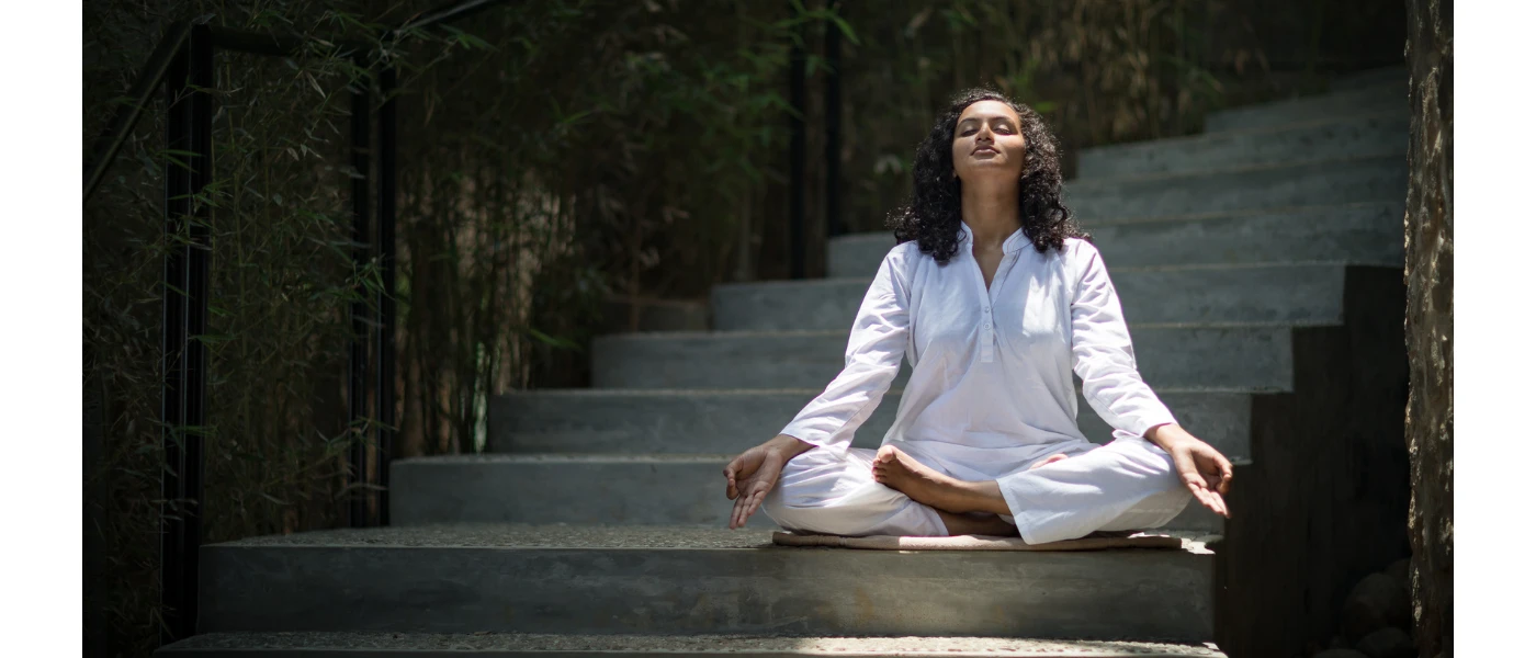 Woman in white sits cross legged with her eyes closed and palms up on a stone staircase