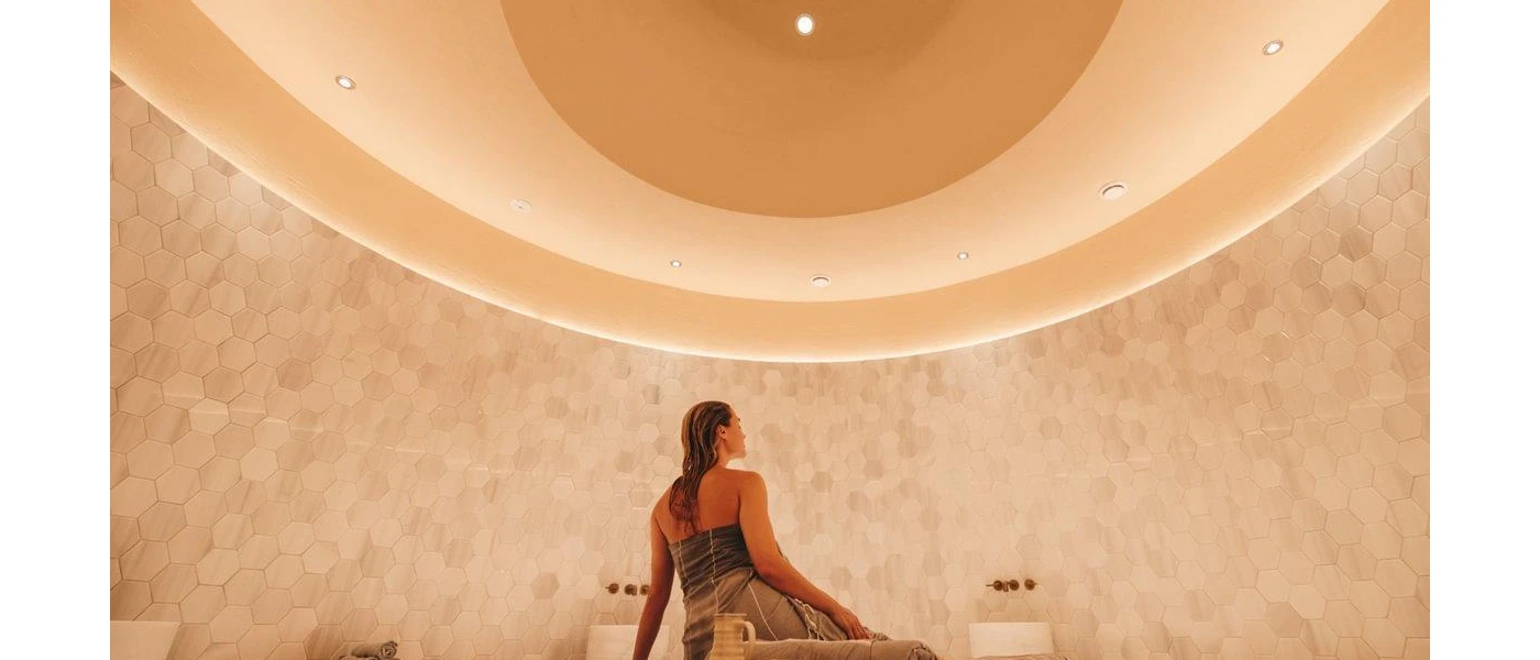 Woman in a towel sitting in the centre of a marble grey-tiled room, gazing up at a spotlighted ceiling