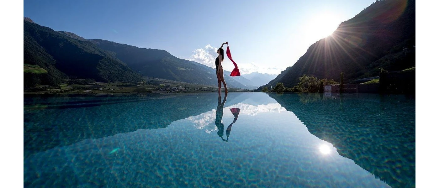 Woman in black swimsuit stands on the edge of a pool holding a red scarf, with the mountains and sun in the background