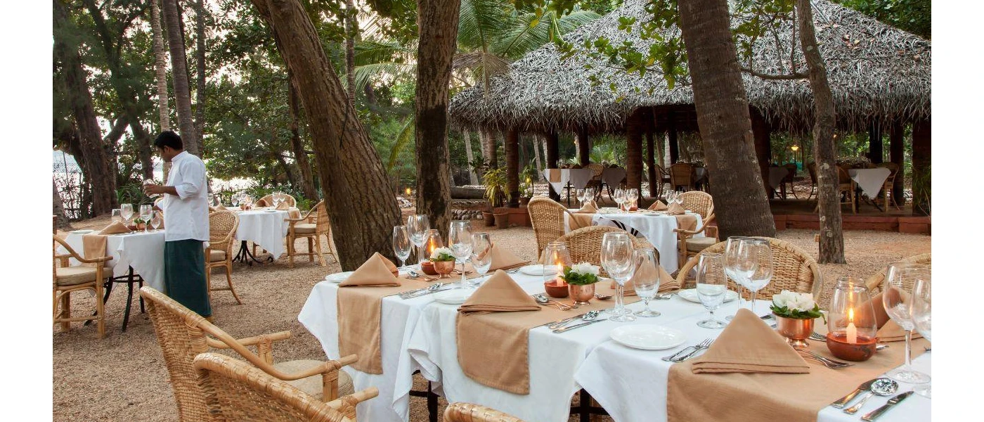 Waiter laying tables with white tablecloths and beige napkins in an outdoor setting surrounded by trees