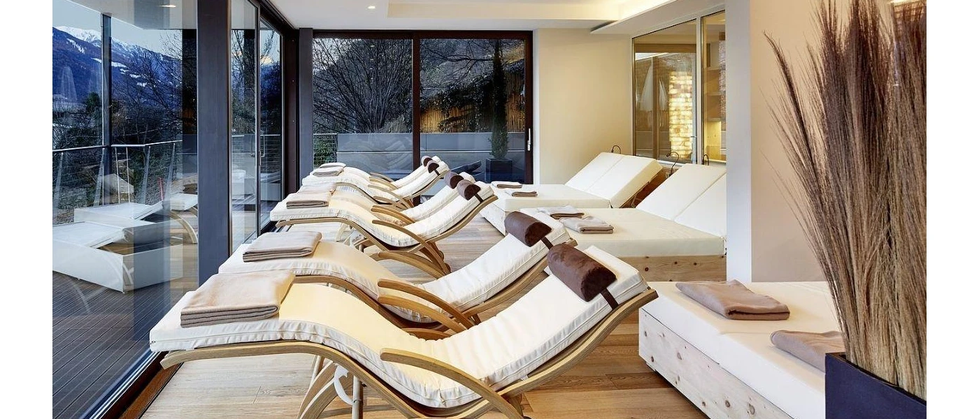Relaxation room with floor-to-ceiling windows, wooden loungers and daybeds with white cushions and towels on top, and pampas grass to one side