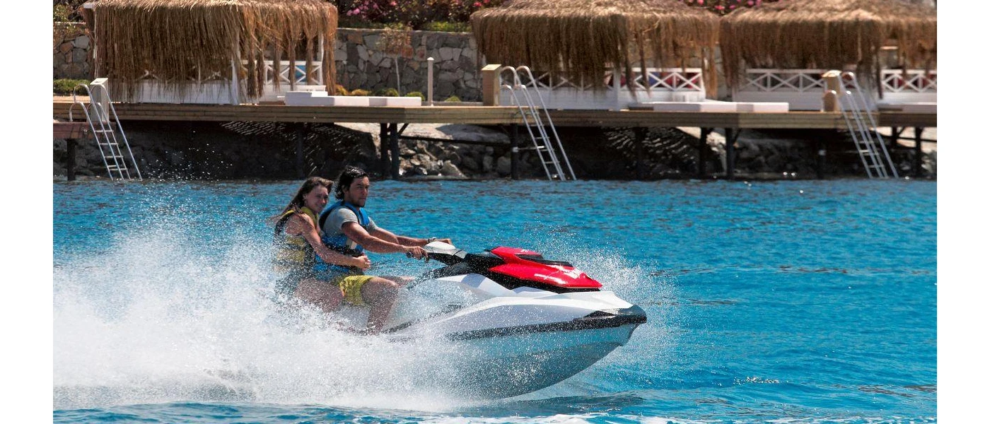 Couple on a jet ski driving past thatched-roof cabanas on the shore