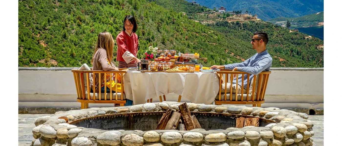 Couple enjoying breakfast in the sunshine overlooking green hills while served by a staff member in a red robe next to a fire pit