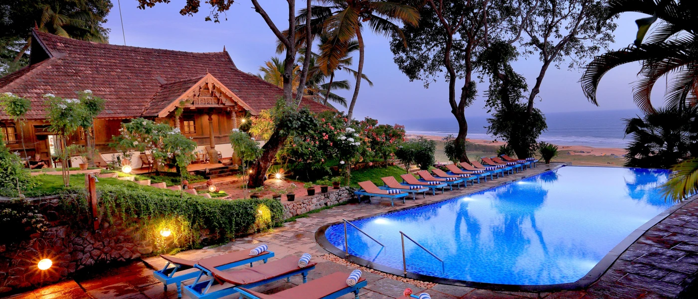 Swimming pool above a golden beach surrounded by orange-cushioned loungers with blue and white striped towels, tropical gardens and neighboured by a traditional Indian building