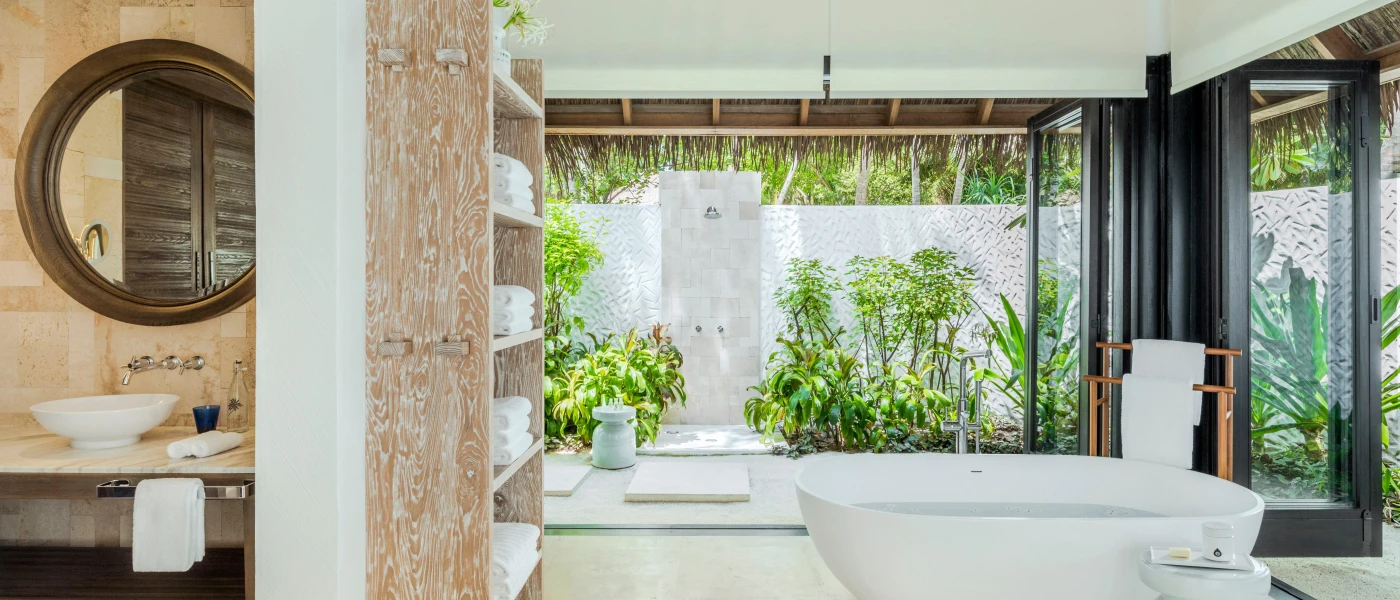 Bathroom with a standalone white tub, shelves of neatly folded white towels, and a floor-to-ceiling window overlooking the greenery beyond