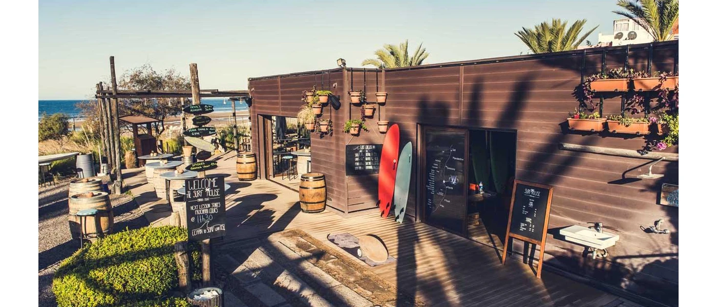 Hut-style building with red and blue surf boards leaning up against it and chalkboards details surf school and timings