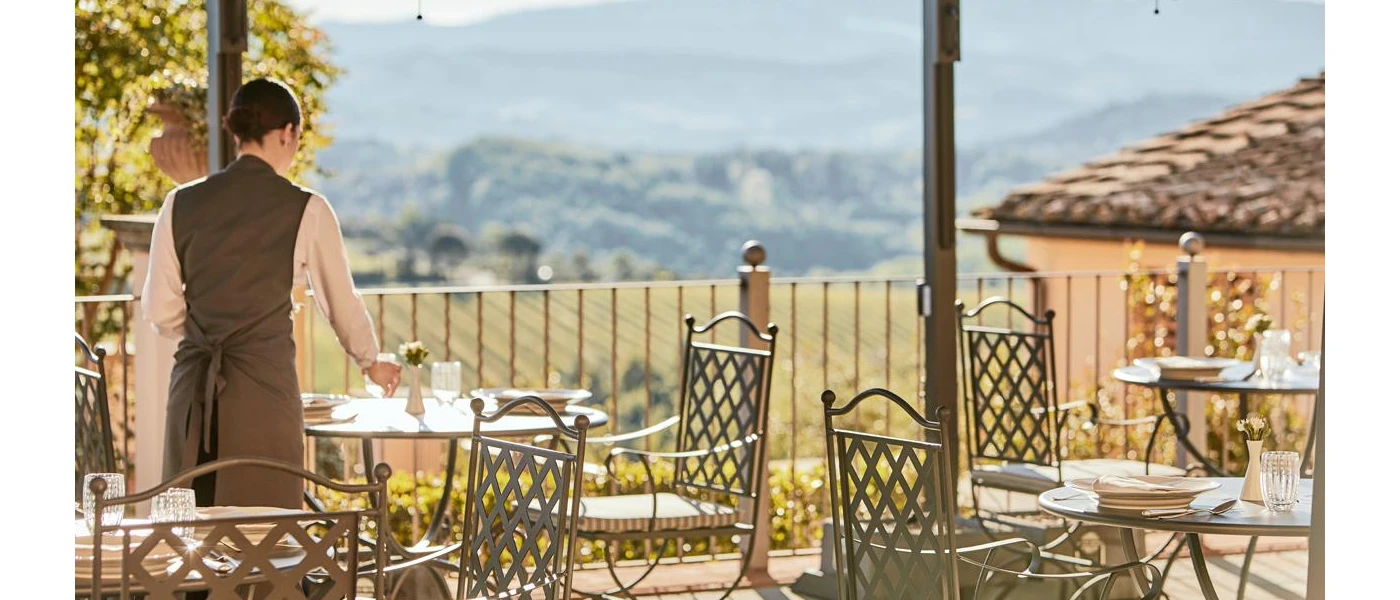 Waitress in a dark waistcoat setting the table on an outdoor terrace with black iron chairs and a view of the Tuscan hills