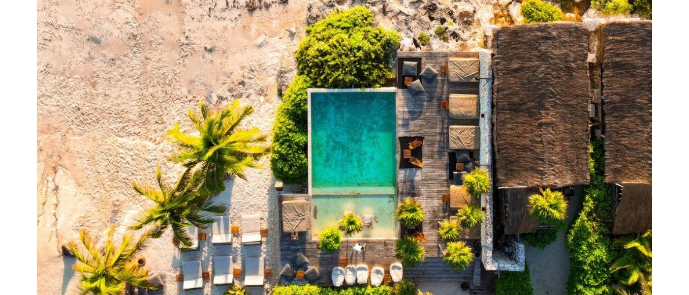 Swimming pool, thatched-roof buildings, loungers and a sandy beach covered in tropical greenery, as seen from above