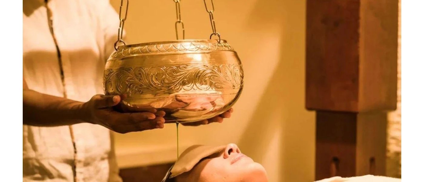 Woman lying down with an eye mask on as a therapist pours oil over her head from a golden bowl