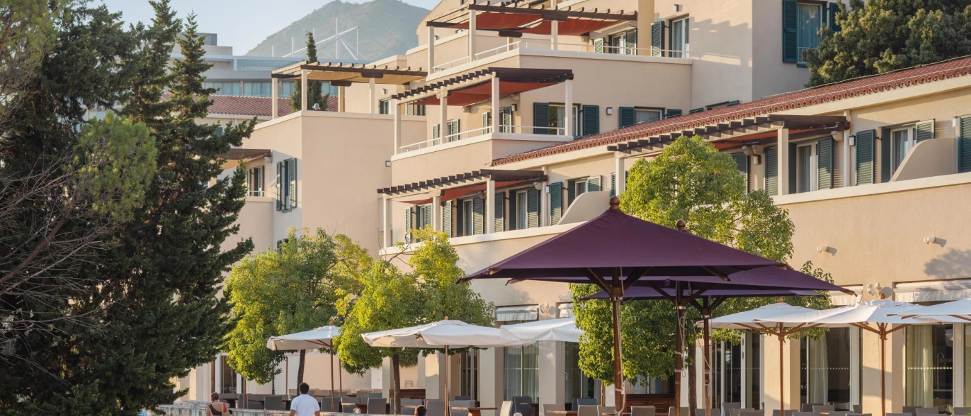 Sand-coloured building fronted by white umbrellas and greenery, as people in summer clothes walk by