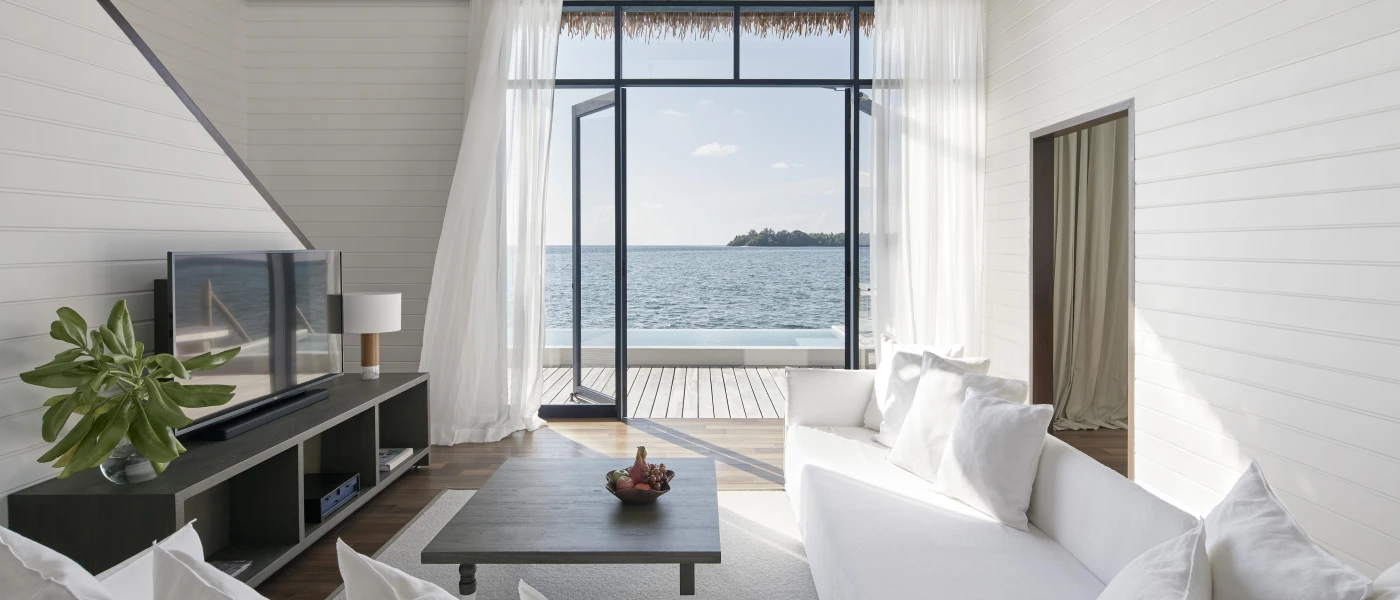 Villa living area with white sofas and walls, a dark-wood coffee table and TV unit and a view of the ocean through open doors