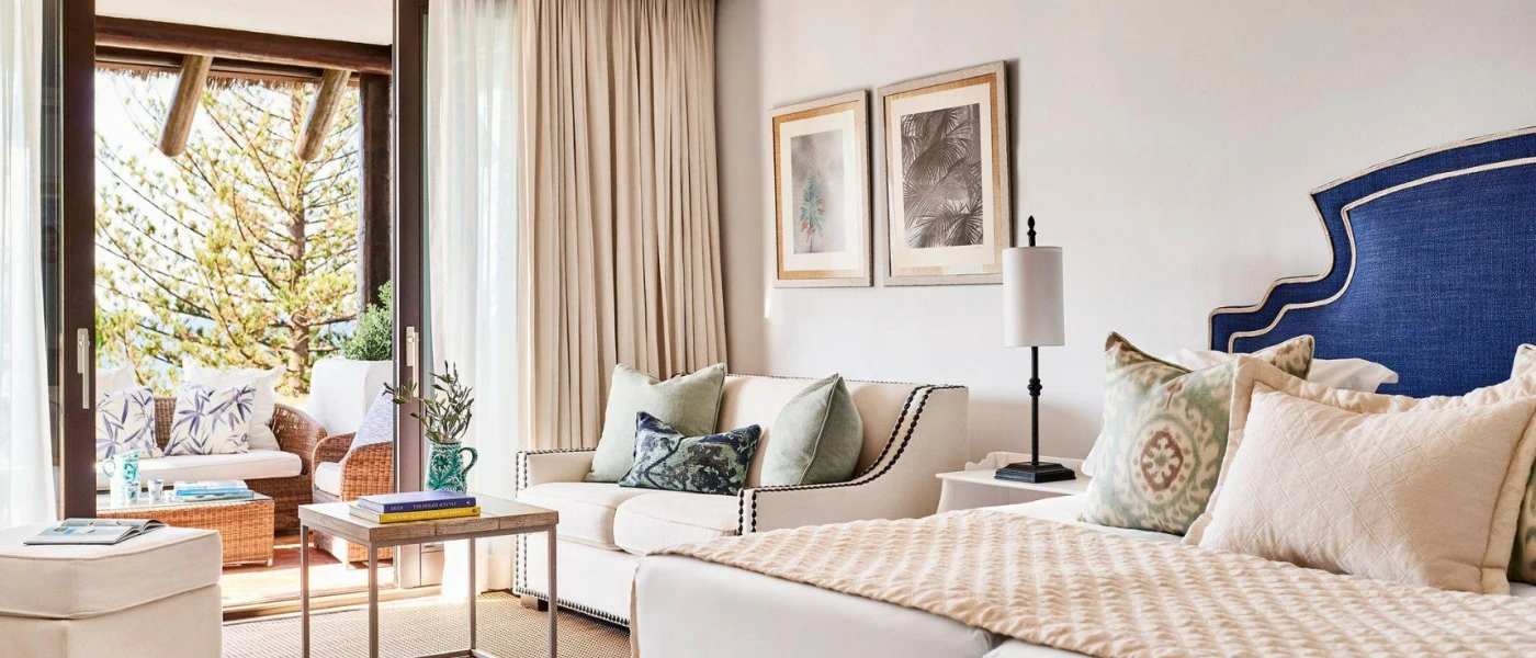 Neutral hotel room with pops of colour in the form of a blue bed headboard, patterned cushions and a balcony with wicker furniture 