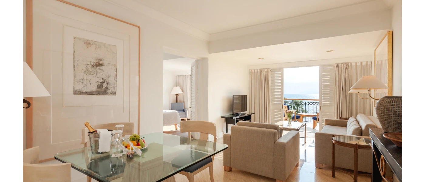Hotel room living and dining area, with a glass dining table, bucket with a bottle and a fruit platter, next to a neutral sofa area and a balcony with a sea view
