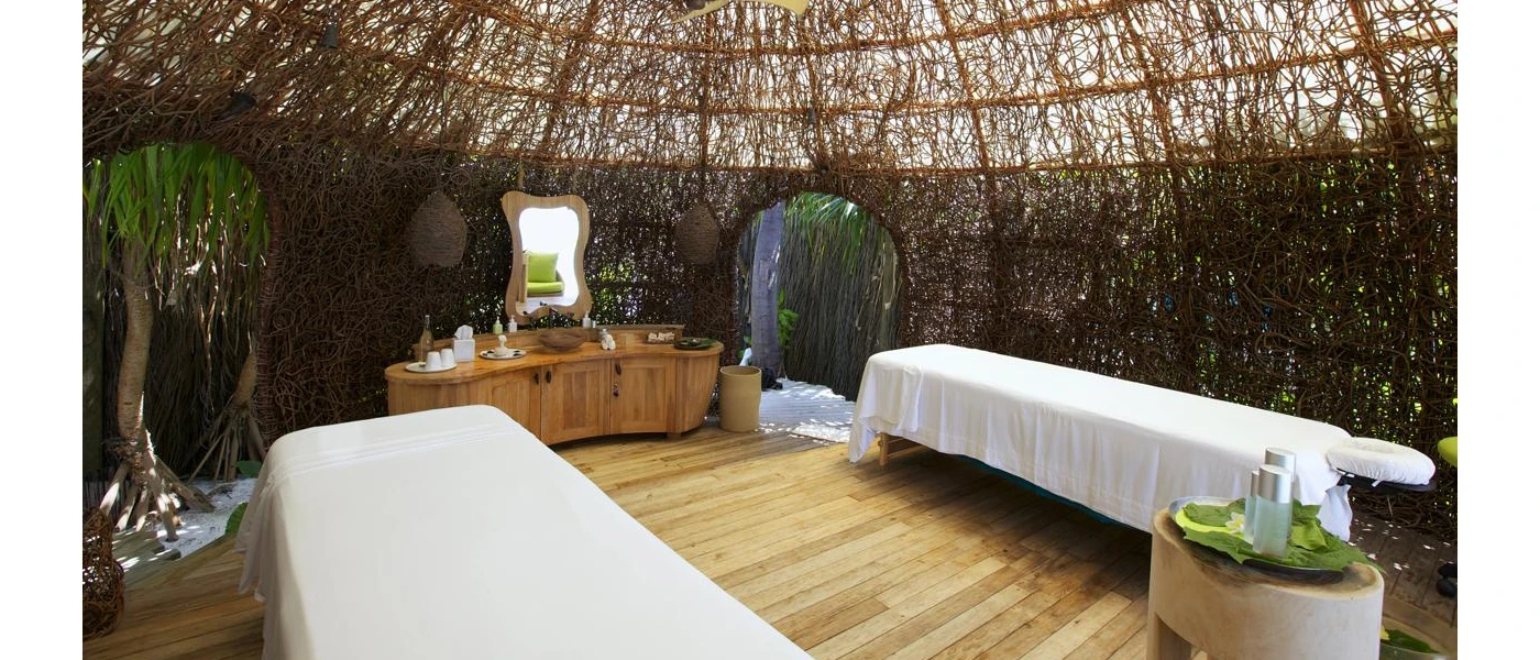 Treatment room made out of a nest-like material, with two massage beds cloaked in white and a wooden vanity unit with a mirror 