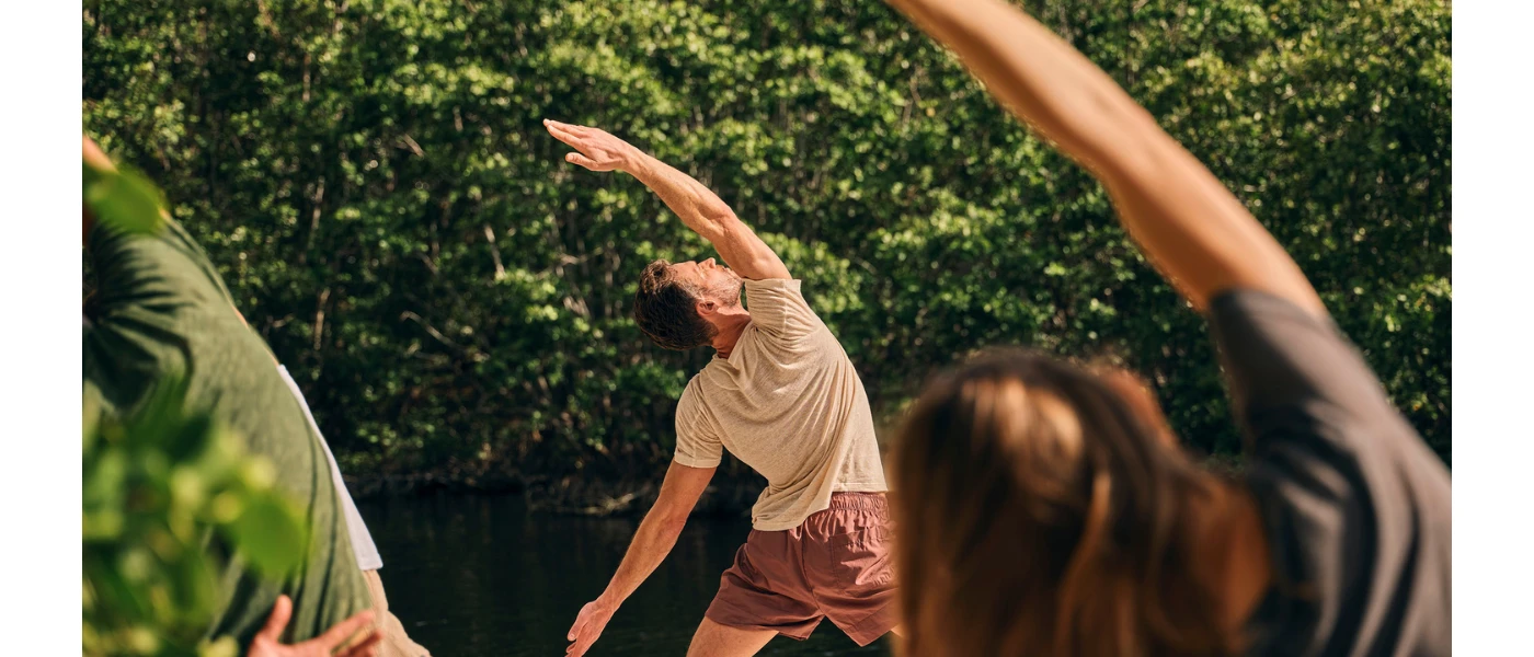A man and woman in active wear stretch over to the left in a sunny garden setting