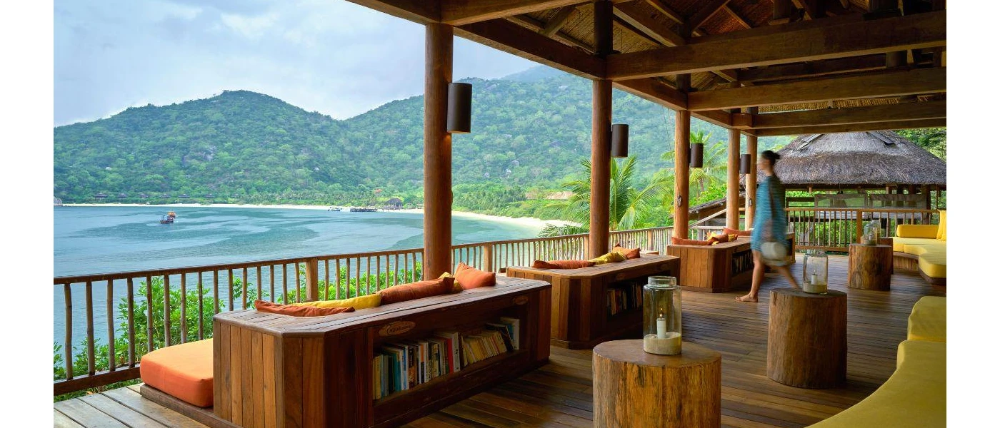 Ocean-facing, open sided pavilion with wooden tables, orange-cushioned sofas and wooden shelves stacked with books