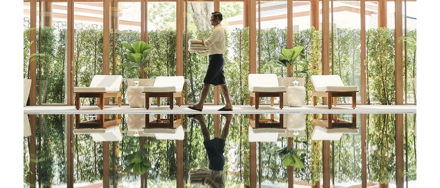 Staff member in dark shorts and a white shirt carries white towels through a room with an indoor pool, ivory-cushioned loungers and floor-to-ceiling windows showcasing tropical greenery