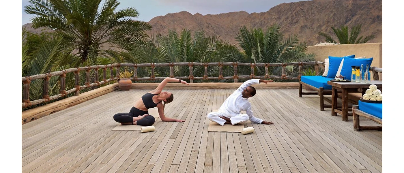 A woman in active wear with a staff instructor in traditional white dress, practicing yoga on a wooden deck surrounded by palm trees and mountains