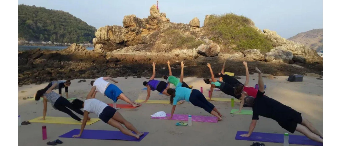 Group in active wear practice yoga on colourful yoga mats next to rocks under a blue sky