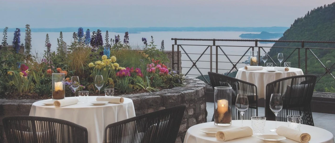 Outdoor terrace with tables with white tablecloths and candles, a flower bed and lake views
