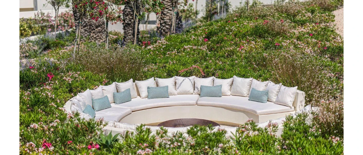 Circular ivory sofa with white and blue cushions fringed by bushes and flowers