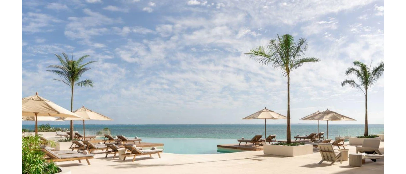 Infinity edge pool in a sunny terrace with tall palm trees, and sleek light-wood loungers with beige cushions