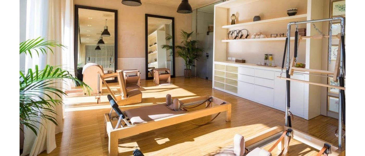 Airy Pilates studio with Reformer machines, full-length black-framed mirrors, glossy green plants, and a white cabinet with ornaments and fruit water