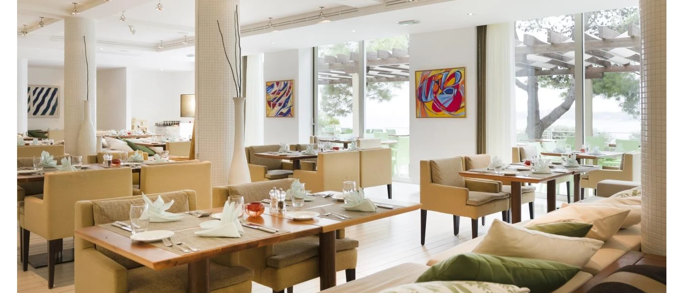 Restaurant with white walls, neutral chairs and wooden tables, plus colourful artwork on the walls, looking out to a terrace with trees