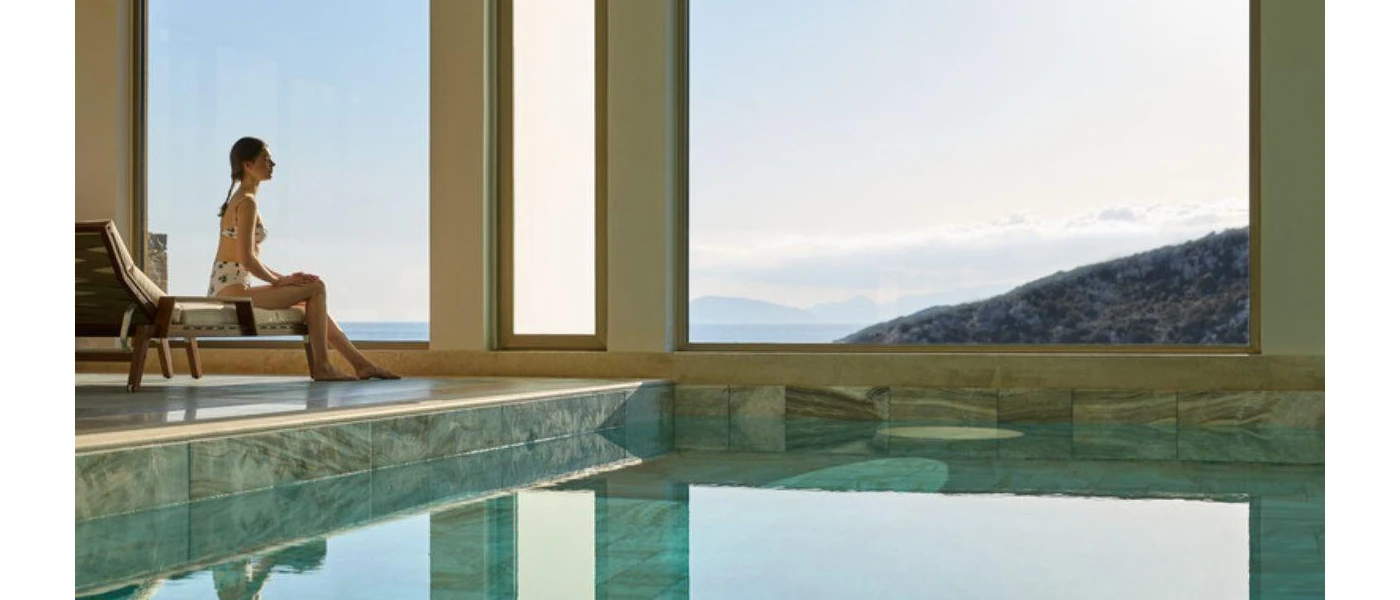 Woman in a printed swimsuit sits by an indoor pool gazing out large windows towards the sea and hillsides
