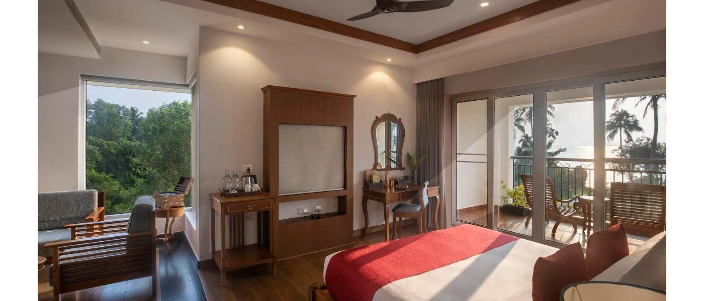 Hotel room with white walls, tiled flooring, white bedding with red cushions and a throw, and dark-wood furnishings, with floor-to-ceiling windows and doors overlooking a balcony