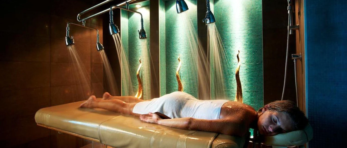 Woman lies on her front on a treatment bed with her eyes closed, as water jets spray onto her in a dark room