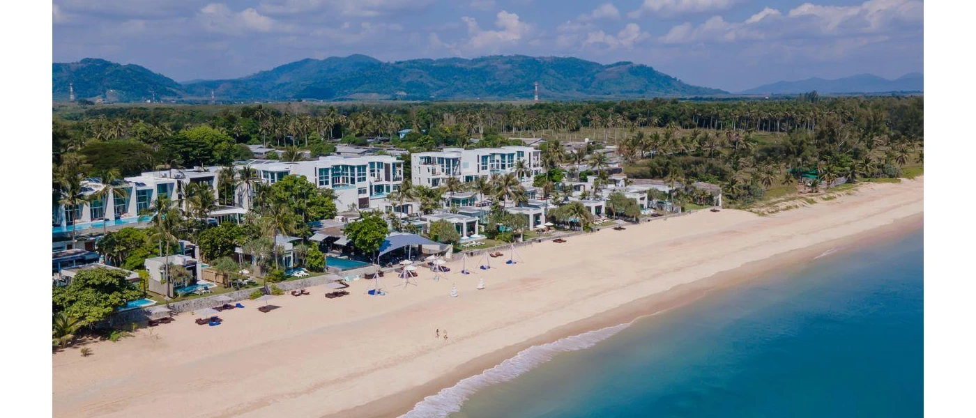 High-up view of white villas and pools on a white sandy beach, fronted by turquoise waves and backed by tropical greenery and mountains