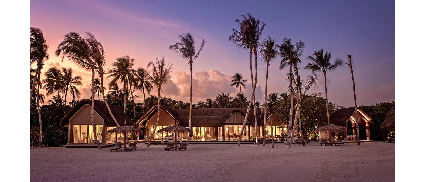 Thatched-roof buildings, tall palm trees and thatched umbrellas on the sand as the sun goes down