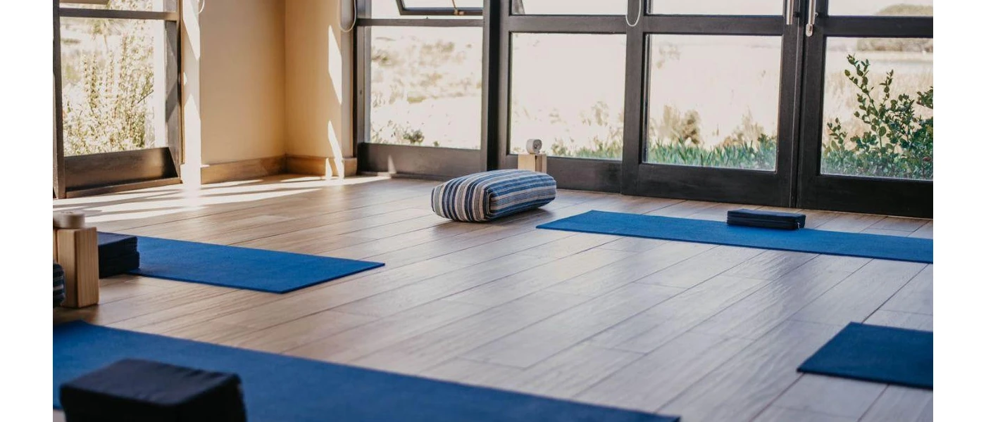 Room with full-length windows and a wooden floor, set up for a class with blue yoga mats and cushions