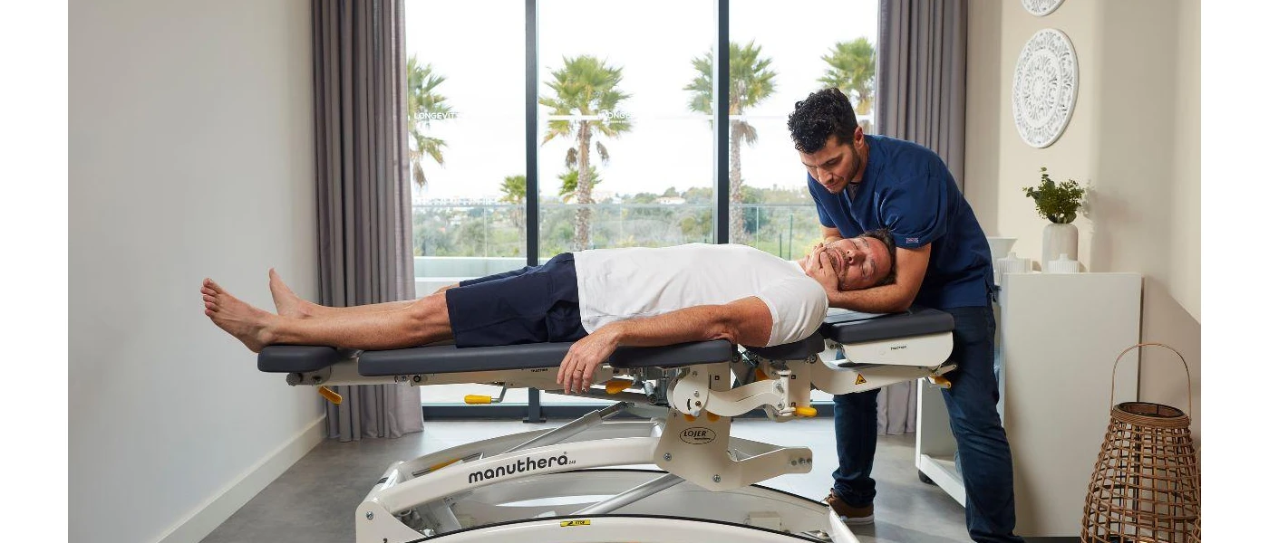 Man in navy shorts and a white t shirt lies on his back on a bed as a therapist in blue uniform treats his neck