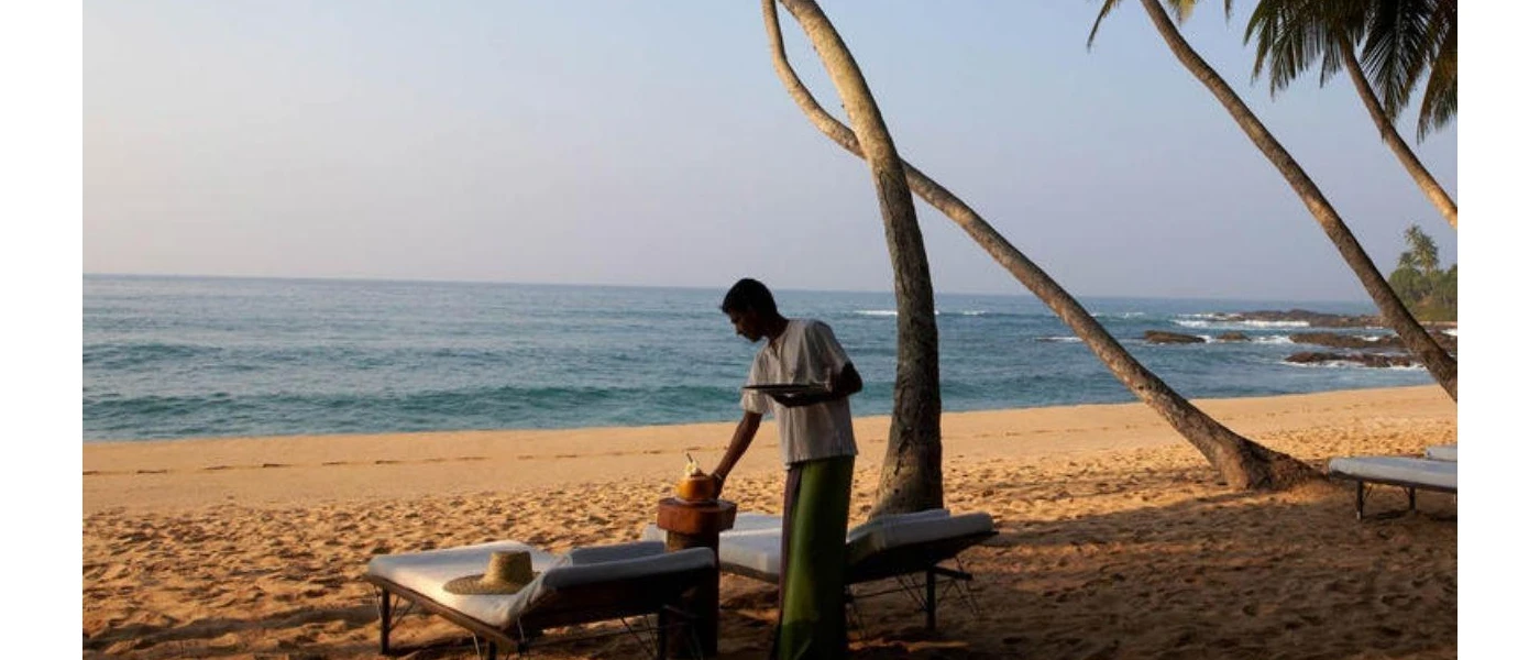 Waiter in traditional dress serving drinks to a table next to the ocean, with loungers and a hat left on one of them