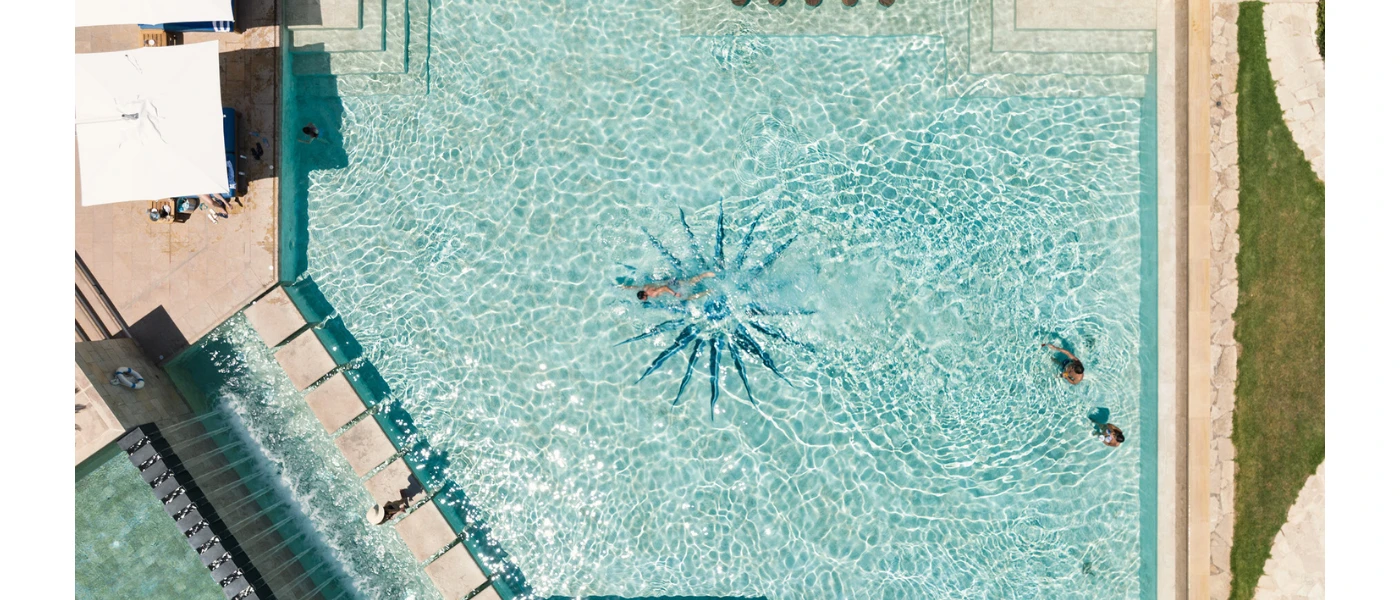 Aerial view of turquoise swimming pool with a star design at its centre, concrete walkway and swimmers enjoying the water in the sunshine
