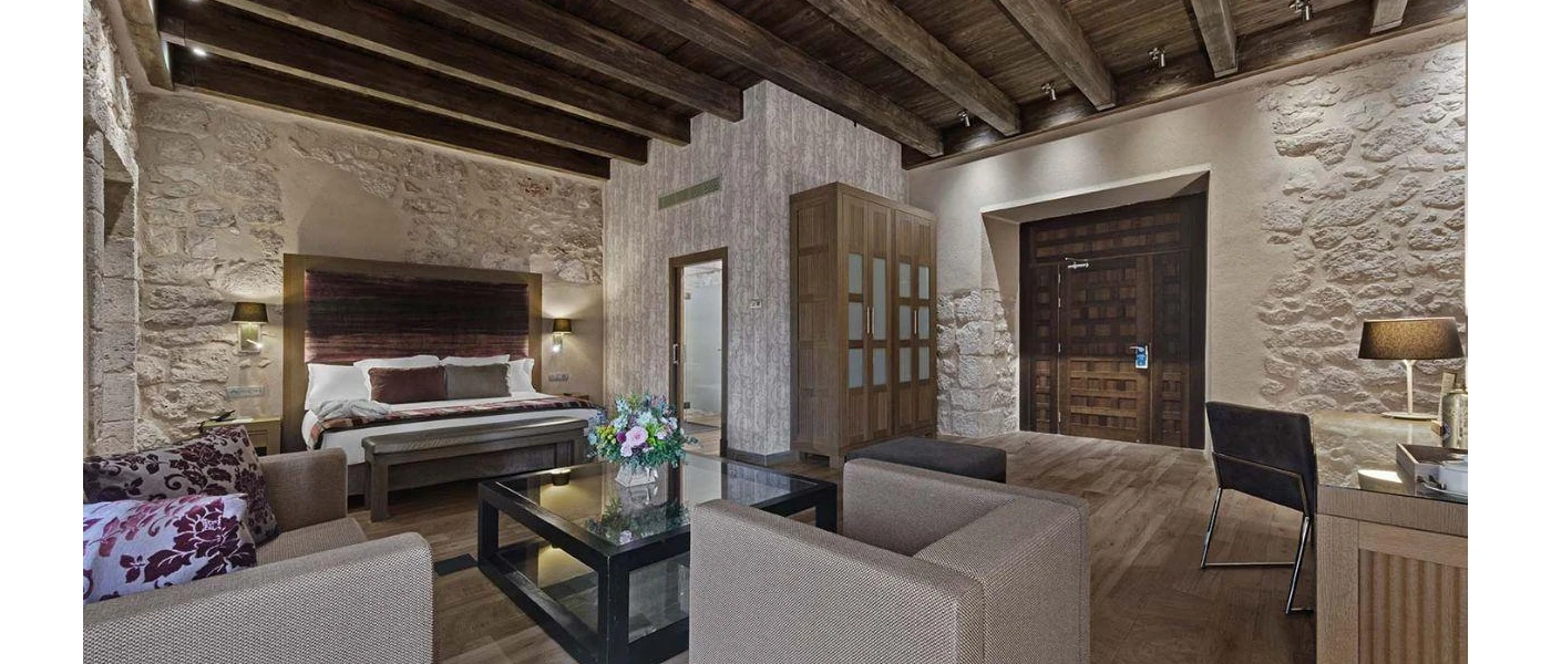 Hotel room with wooden ceiling beams and stone walls, a bed with white bedding and brown and grey cushions, a seating area with a glass coffee table, and a desk area with a lamp