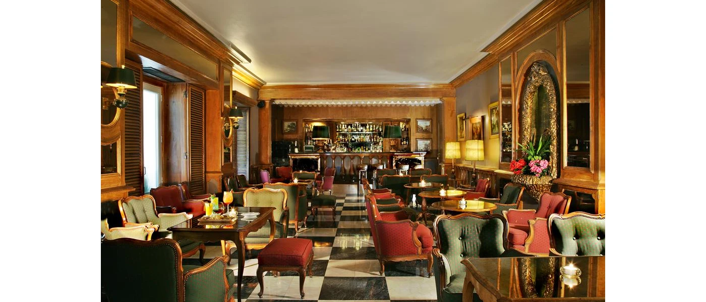 Glamorous traditional bar, with wood-panelled walls, a glossy black and white tiled floor, and gilded red and green armchairs