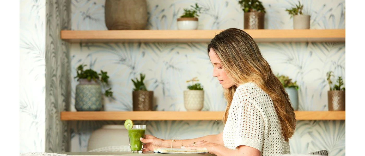 Woman in a white knitted shirt sits with a green smoothie with a kiwi garnish in a room with wooden shelves full of potted green plants