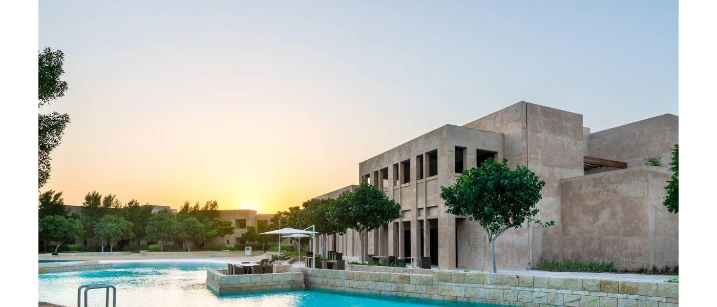 Greenery surrounds a swimming pool under a darkening sky, in front of a sand-coloured building with pillars