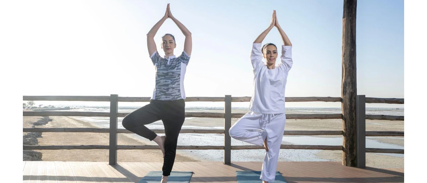 Two women in loose clothing in a yoga pose on a mat on a sunny terrace next to a sandy beach