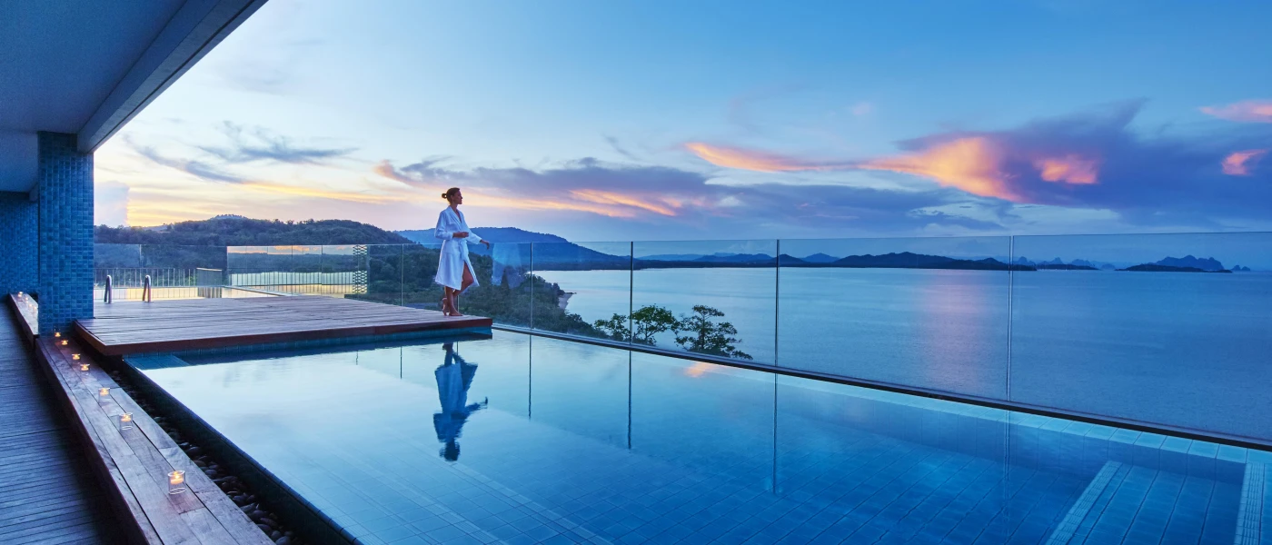 Woman in a white robe gazes out to sea from a glass-fronted balcony, with a private pool and wooden decking scattered with tealight candles
