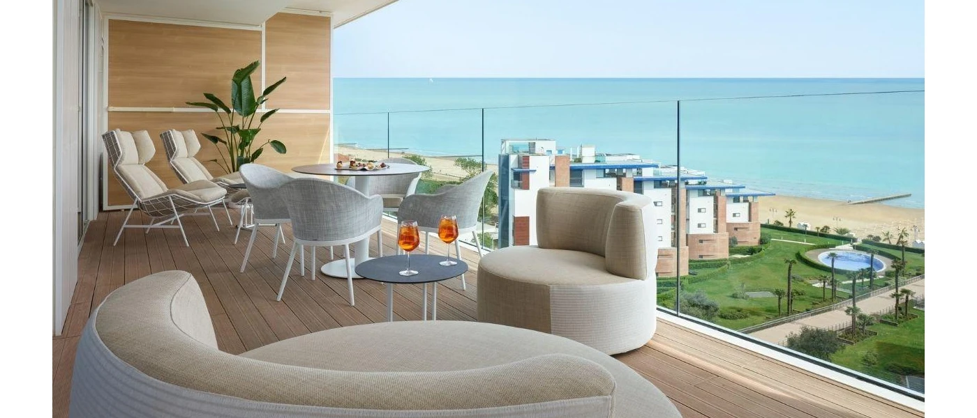 Glass-fronted balcony overlooking gardens and the sea, with neutral chairs and loungers and a table with aperol spritz