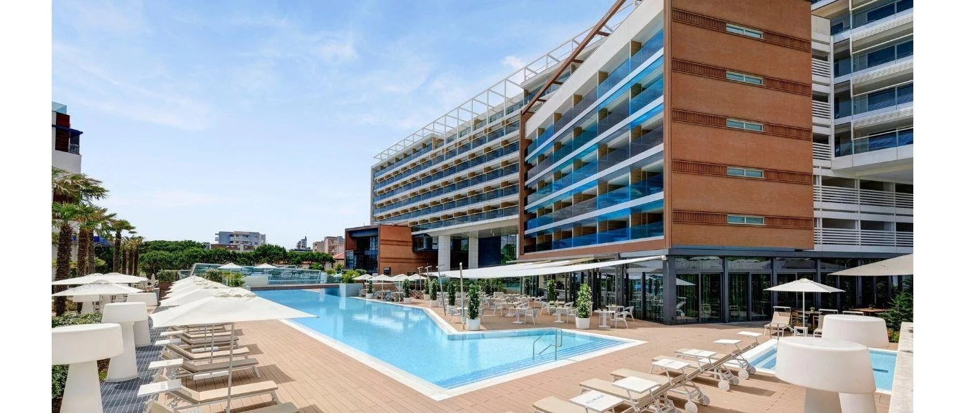 Multi-storey glass fronted hotel with an outdoor pool surrounded by white parasols and loungers