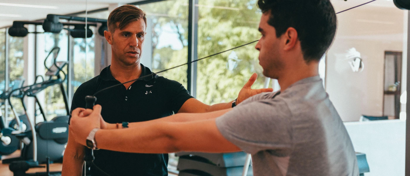 Man in a grey t shirts works out on a weight machine as an instructor in a black polo shirt watches on