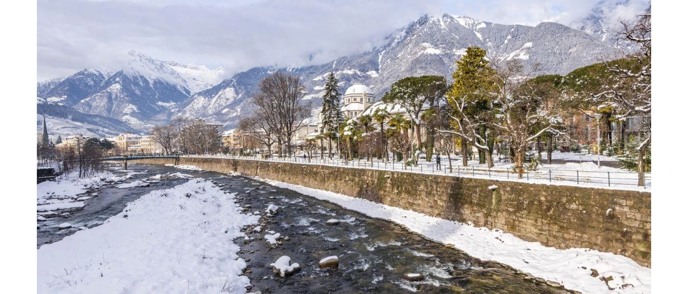 A frozen river under snowy peaks, with a tree-lined promenade and historic architecture