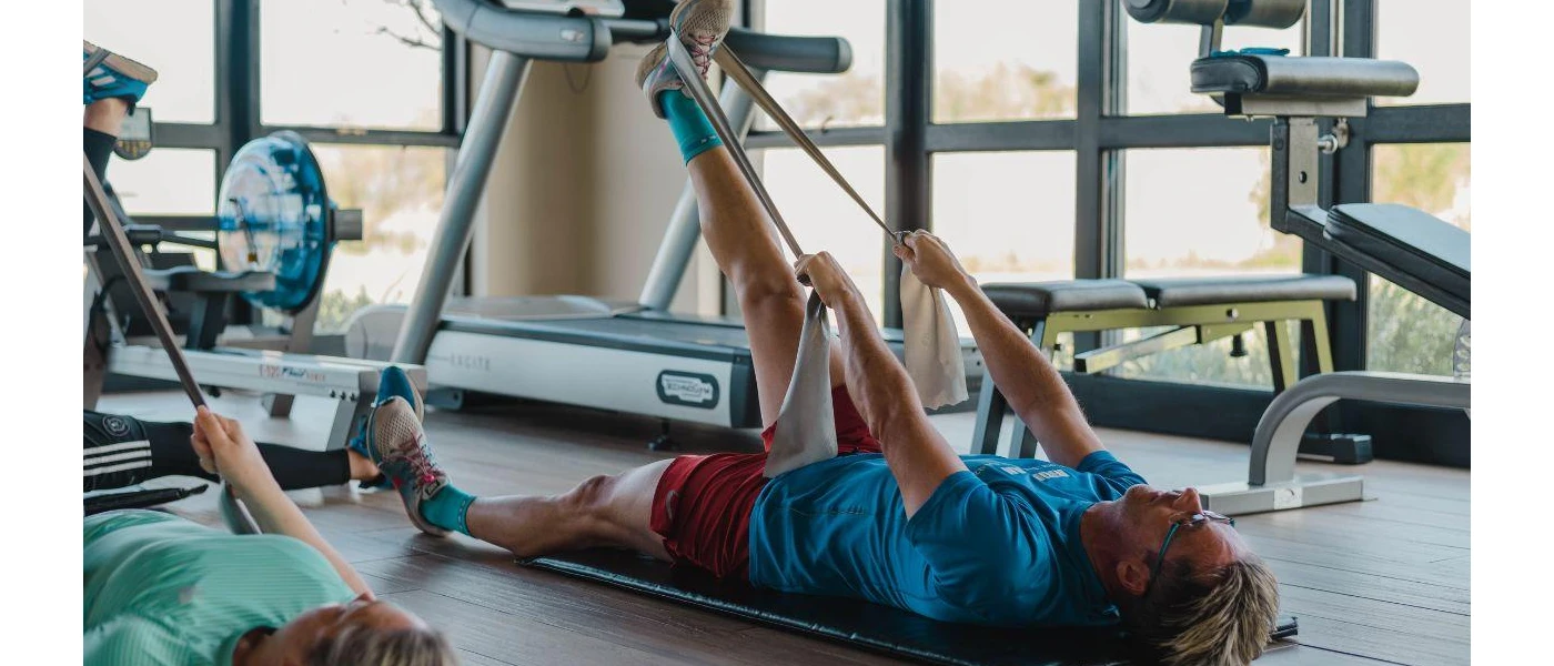 Two people in active wear lying down in a gym as they use resistance bands on one leg, while wearing equipment that measures performance